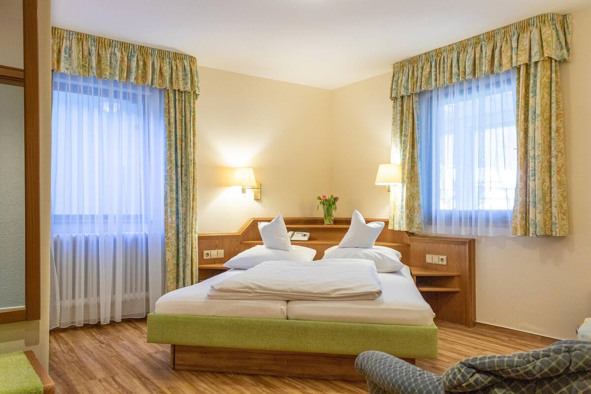 A hotel room with a large bed, two bedside tables, a green bench, and curtains.