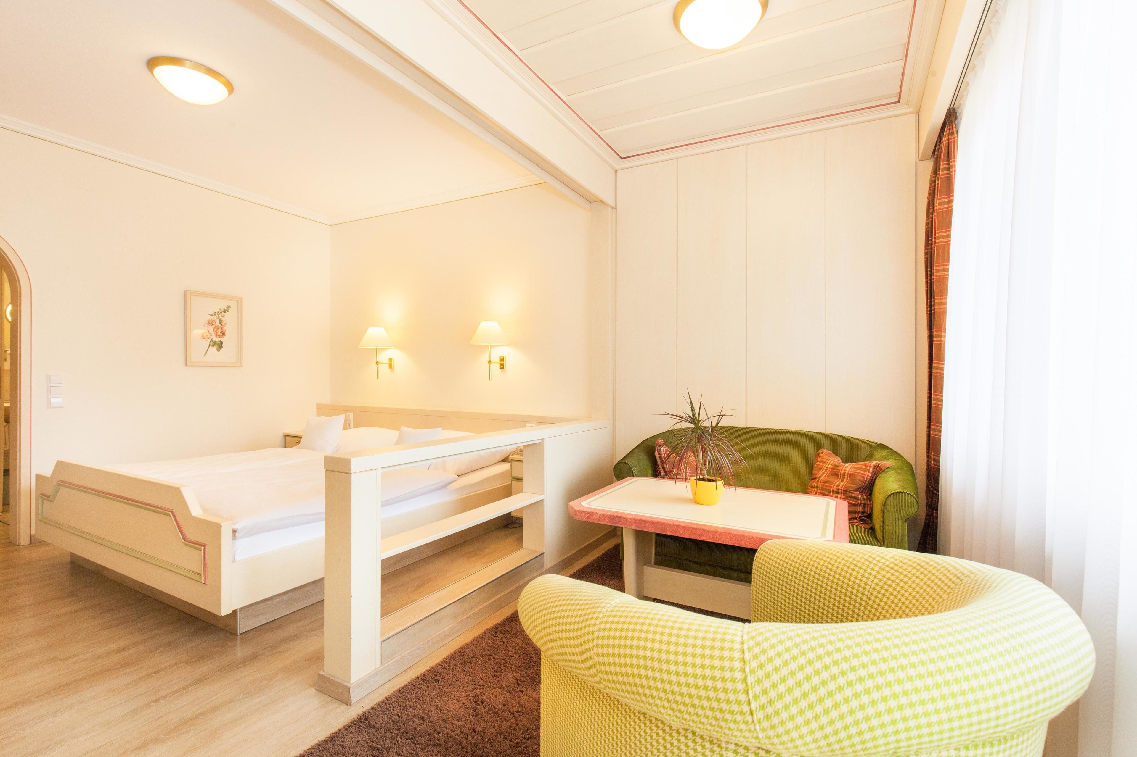 Una habitación con cama, una mesita con planta, una silla y una ventana con cortinas.