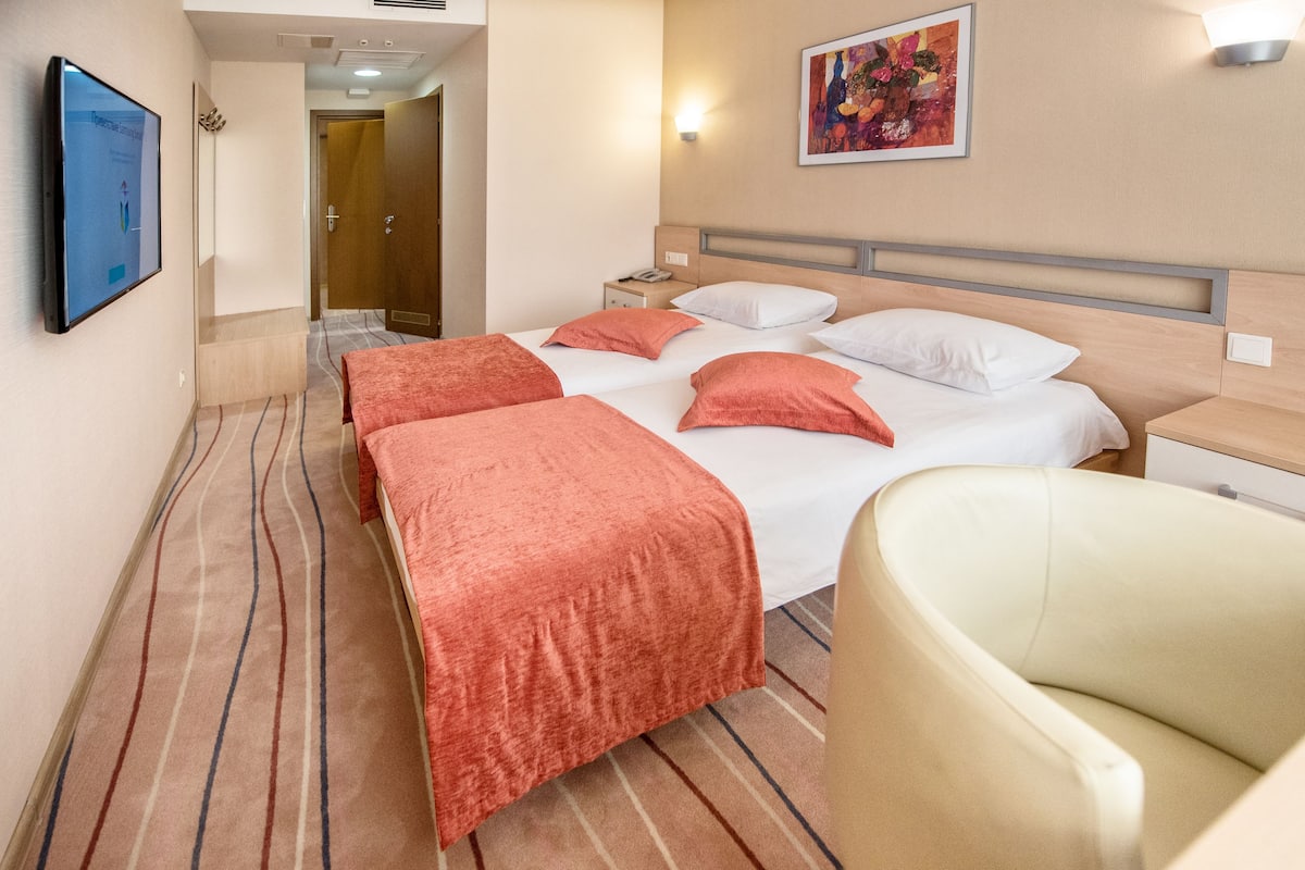 A hotel room with a bed, a chair, a bedside table, a lamp, and a painting on the wall.