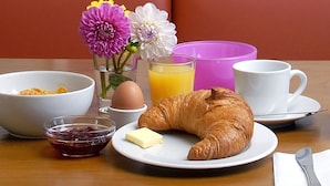 Une table dressée pour le petit-déjeuner avec un croissant, du beurre, de la confiture, un verre de jus d’orange, une tasse de café et un bol de céréales.
