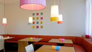 Un espace repas avec des banquettes et des tables, des suspensions colorées et un mur orné d’une grille de photos carrées.