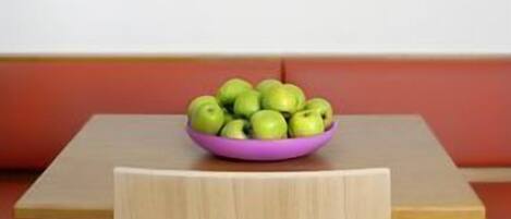 Un comedor con una mesa de madera, un tazón púrpura con manzanas verdes y una lámpara colgante blanca con pantalla azul.