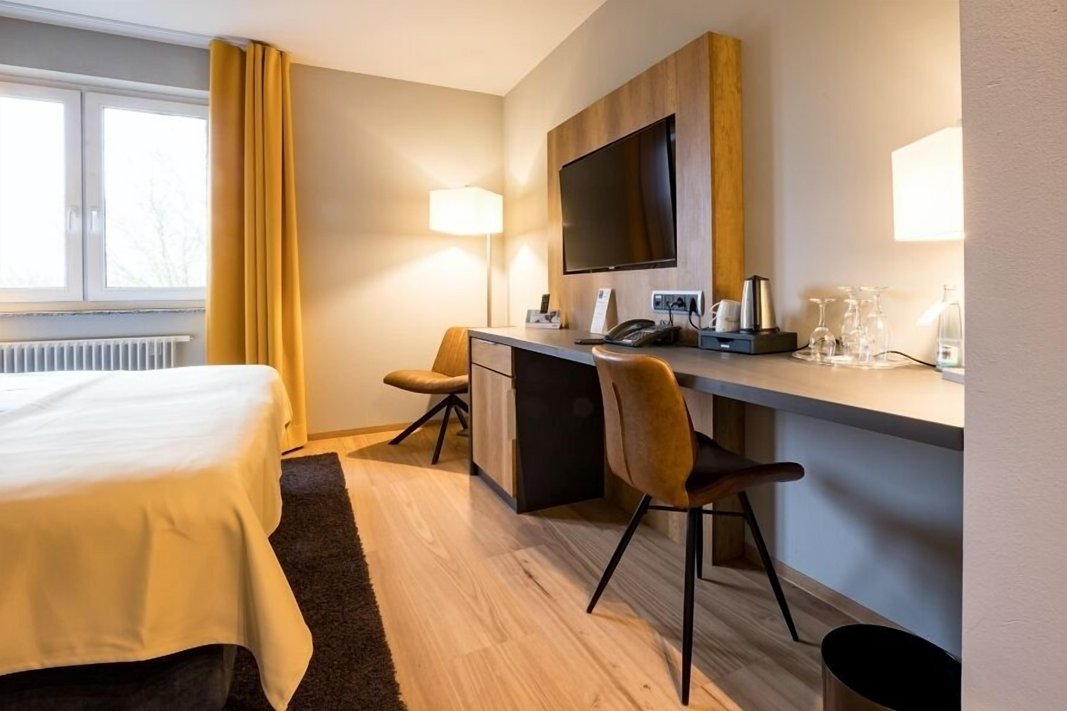 A hotel room with a bed, a desk with a chair, a television, and a window with curtains.