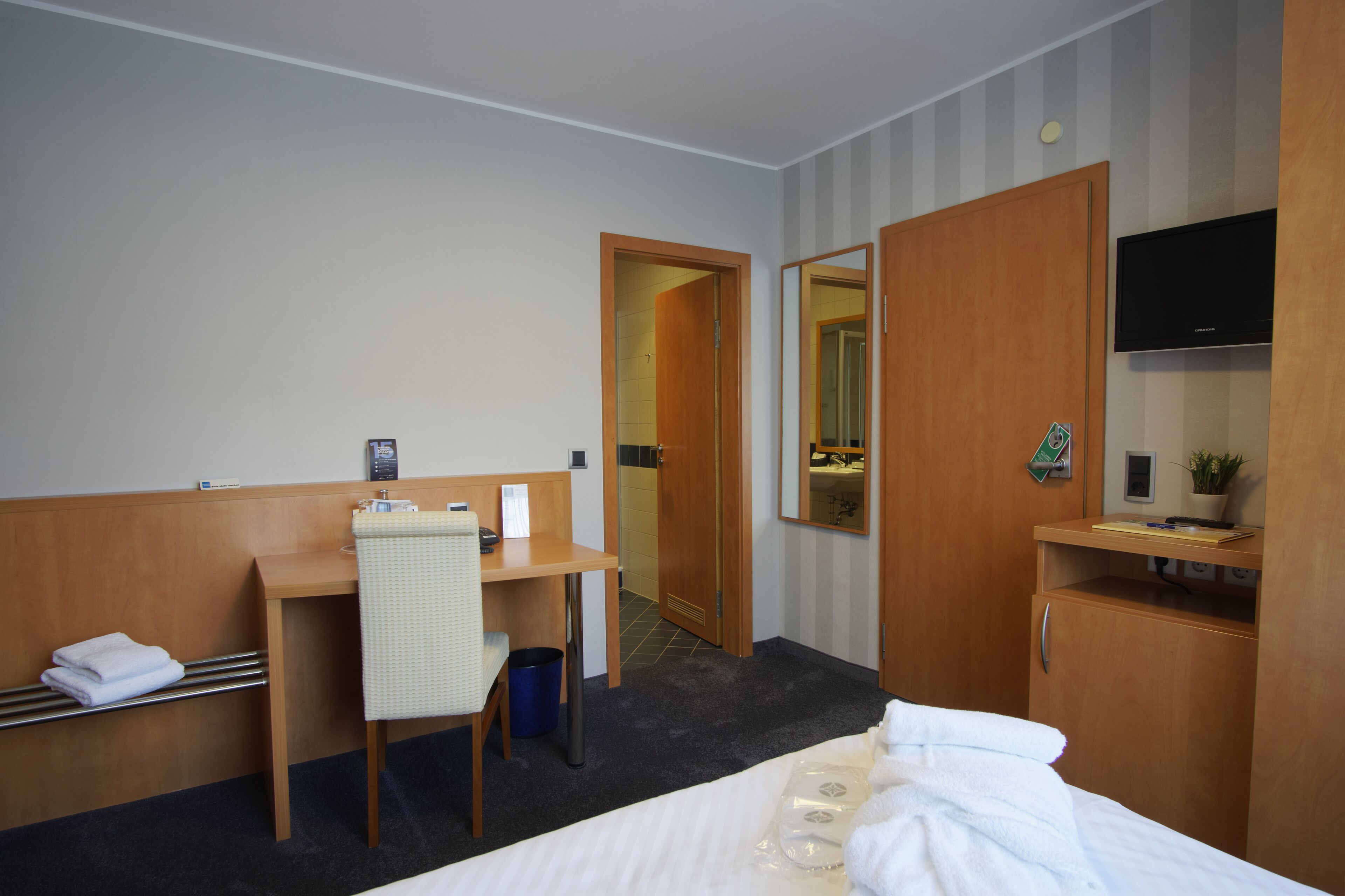 Quarto de hotel com cama, escrivaninha, cadeira, televisão e guarda-roupa.