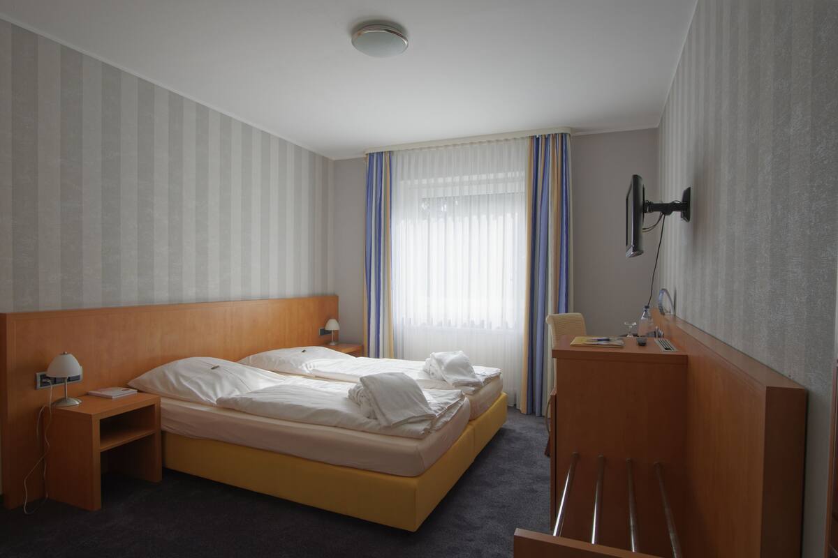 Ein Hotelzimmer mit einem Doppelbett, einem Holzkopfstück, einem Nachttisch mit Lampe, einem Schreibtisch mit Stuhl, einem an der Wand befestigten Flachbildfernseher und einem Fenster mit Vorhängen.