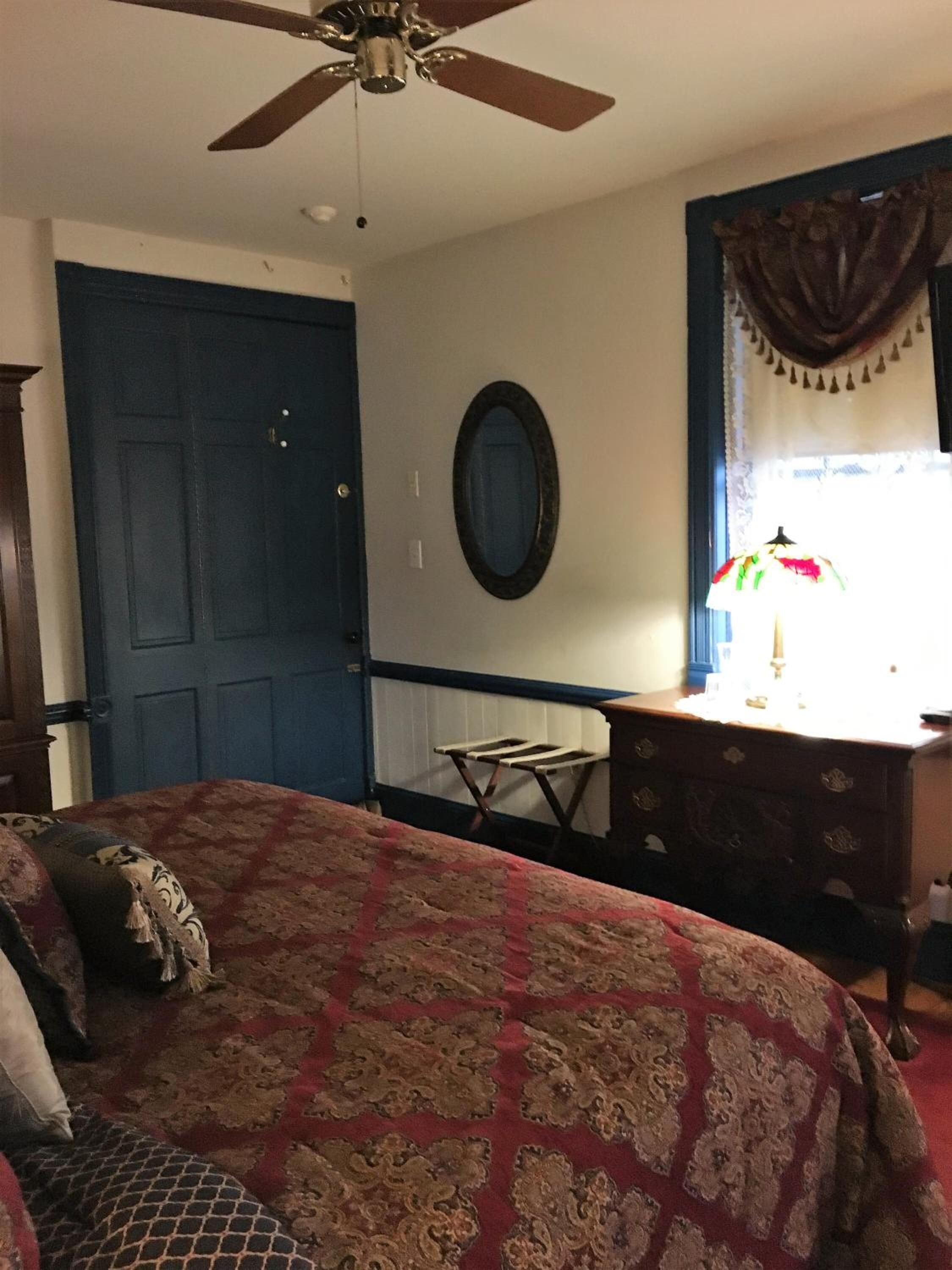 Une chambre à coucher comprenant un lit, une commode, un ventilateur de plafond, une porte et une fenêtre avec des rideaux.