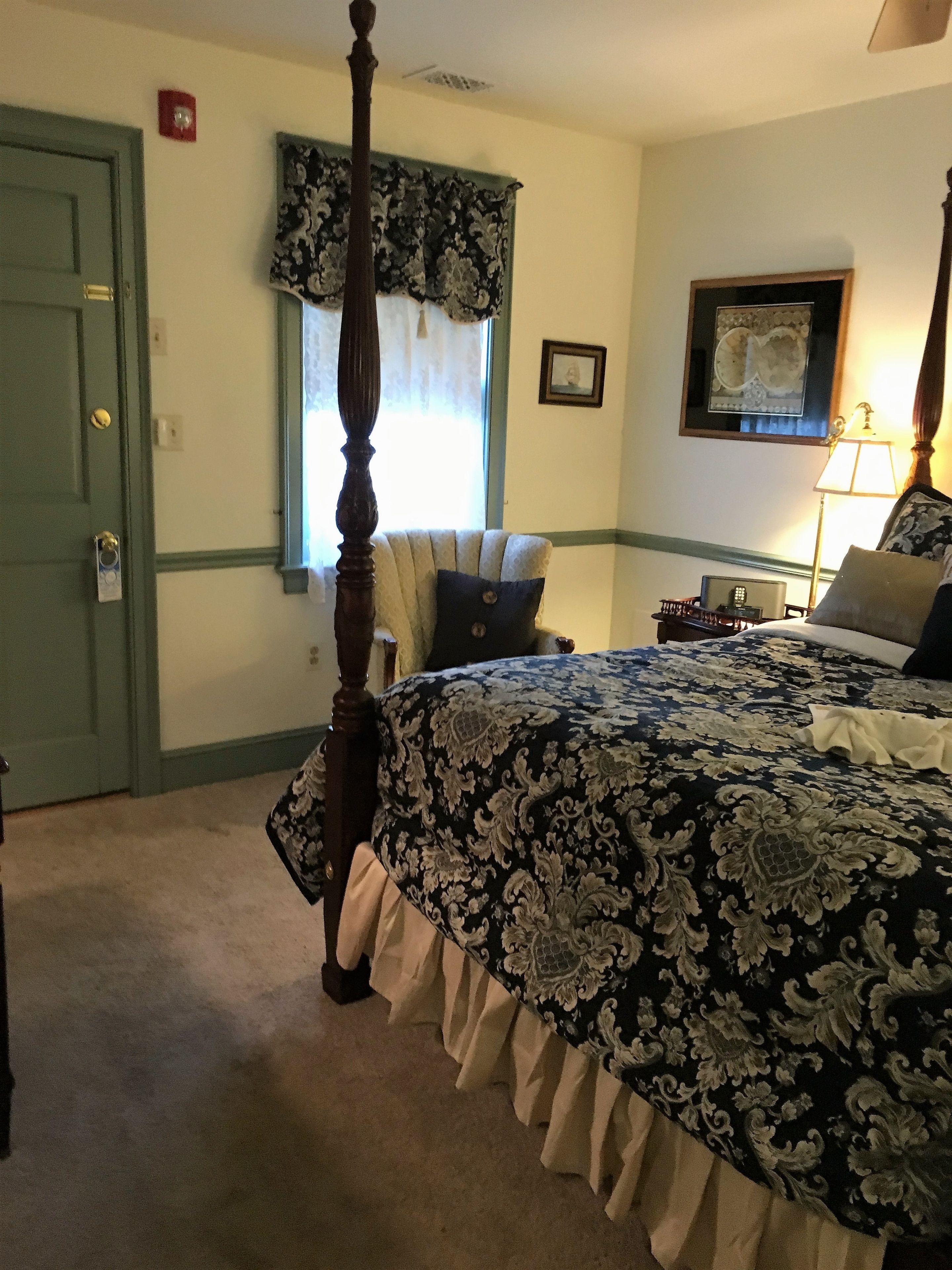 A bedroom with a four-poster bed, a nightstand, a lamp, and a window with curtains.