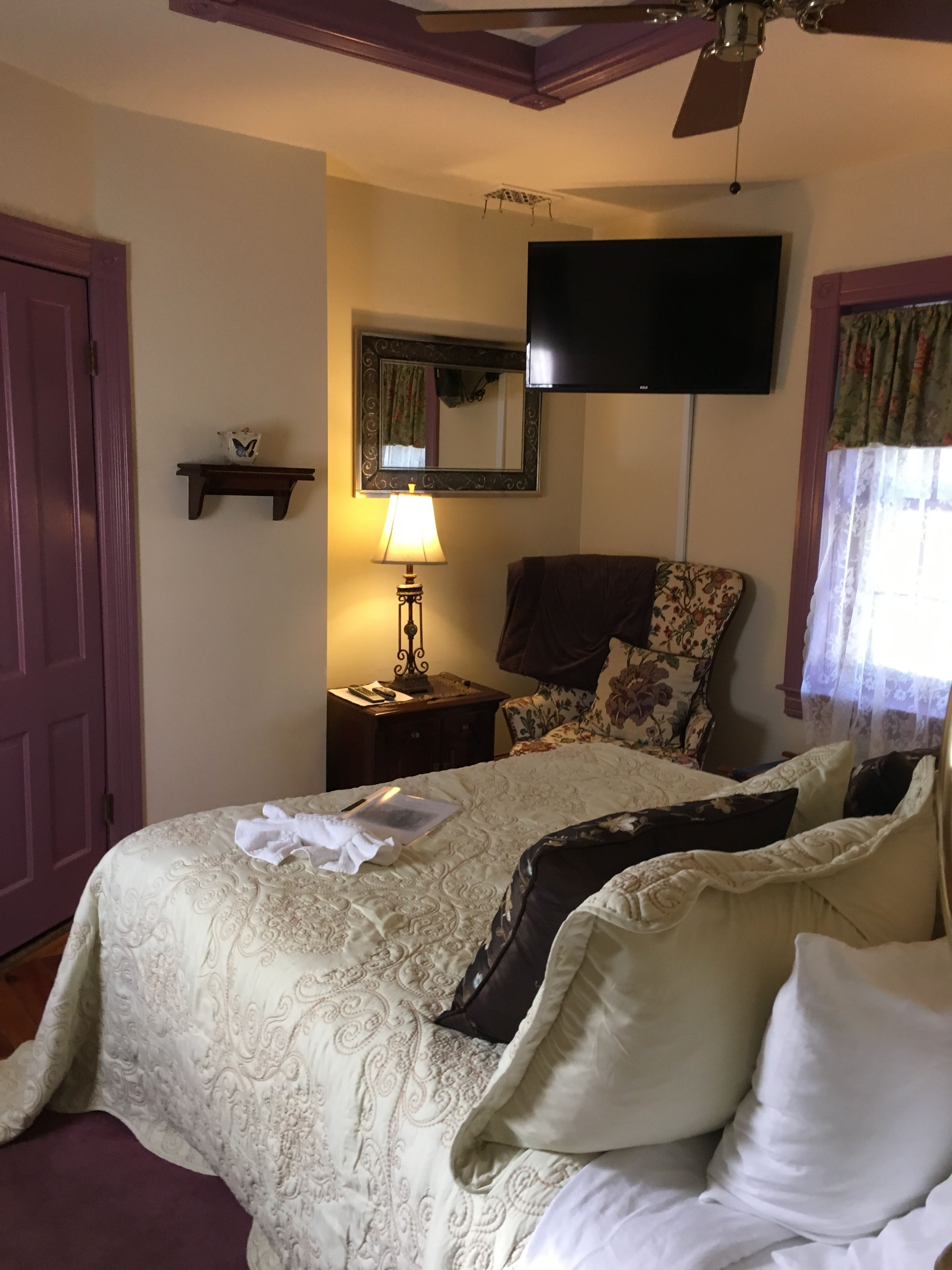 Un dormitorio con una cama, una silla, un televisor, una lámpara y una estantería con un objeto decorativo.