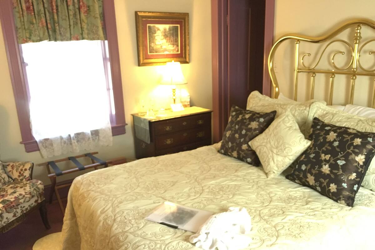 A neatly made bed with floral-patterned pillows, a bedside table with a lamp, and a chair with a floral cover.