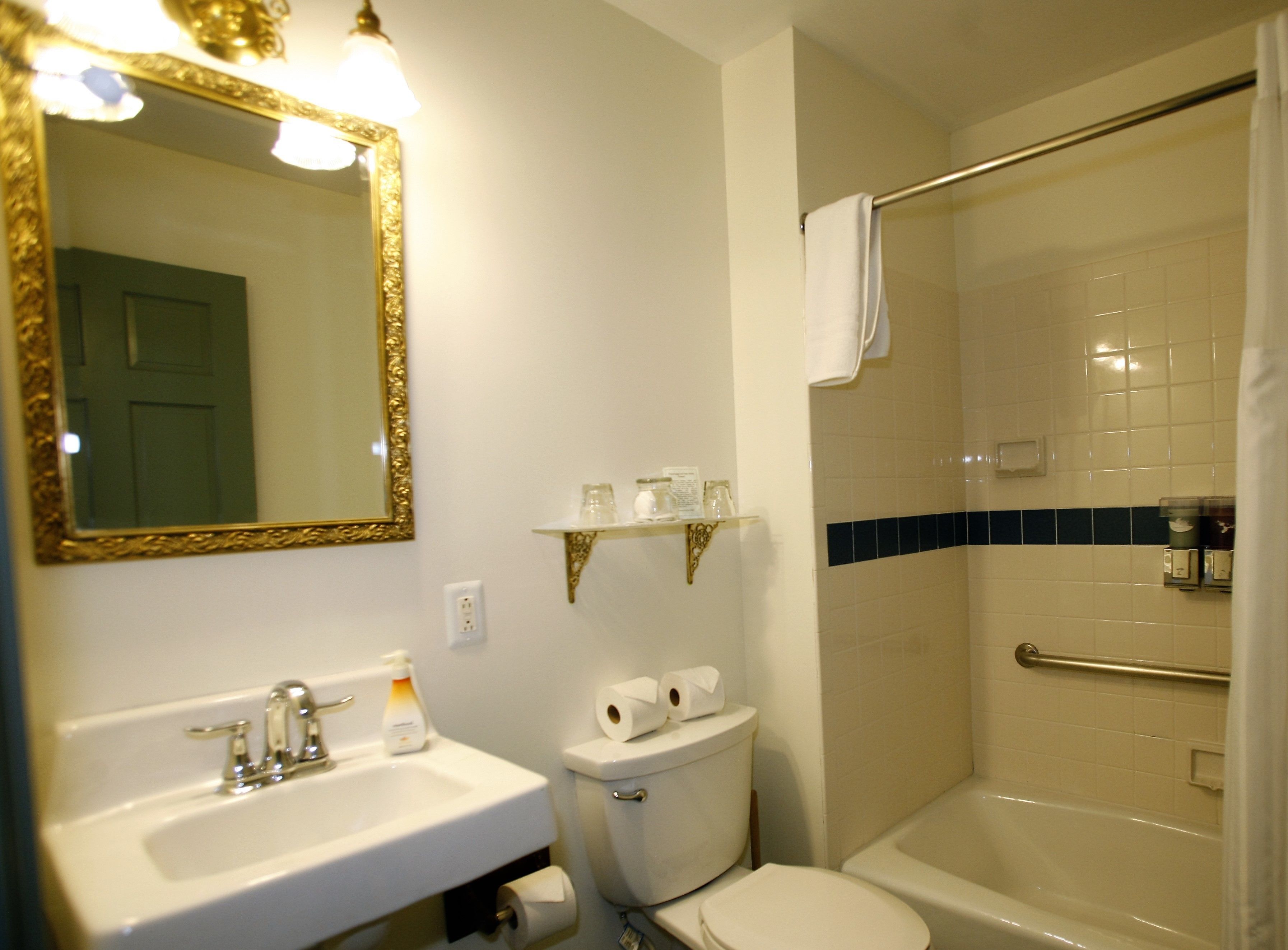 Un baño con lavamanos, inodoro y bañera. Hay un espejo con marco dorado y un estante sobre el inodoro.