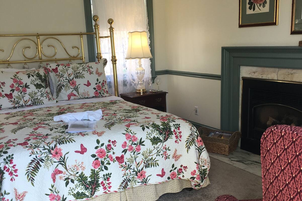 A bedroom with a large bed, a nightstand, a lamp, a chair, and a fireplace.