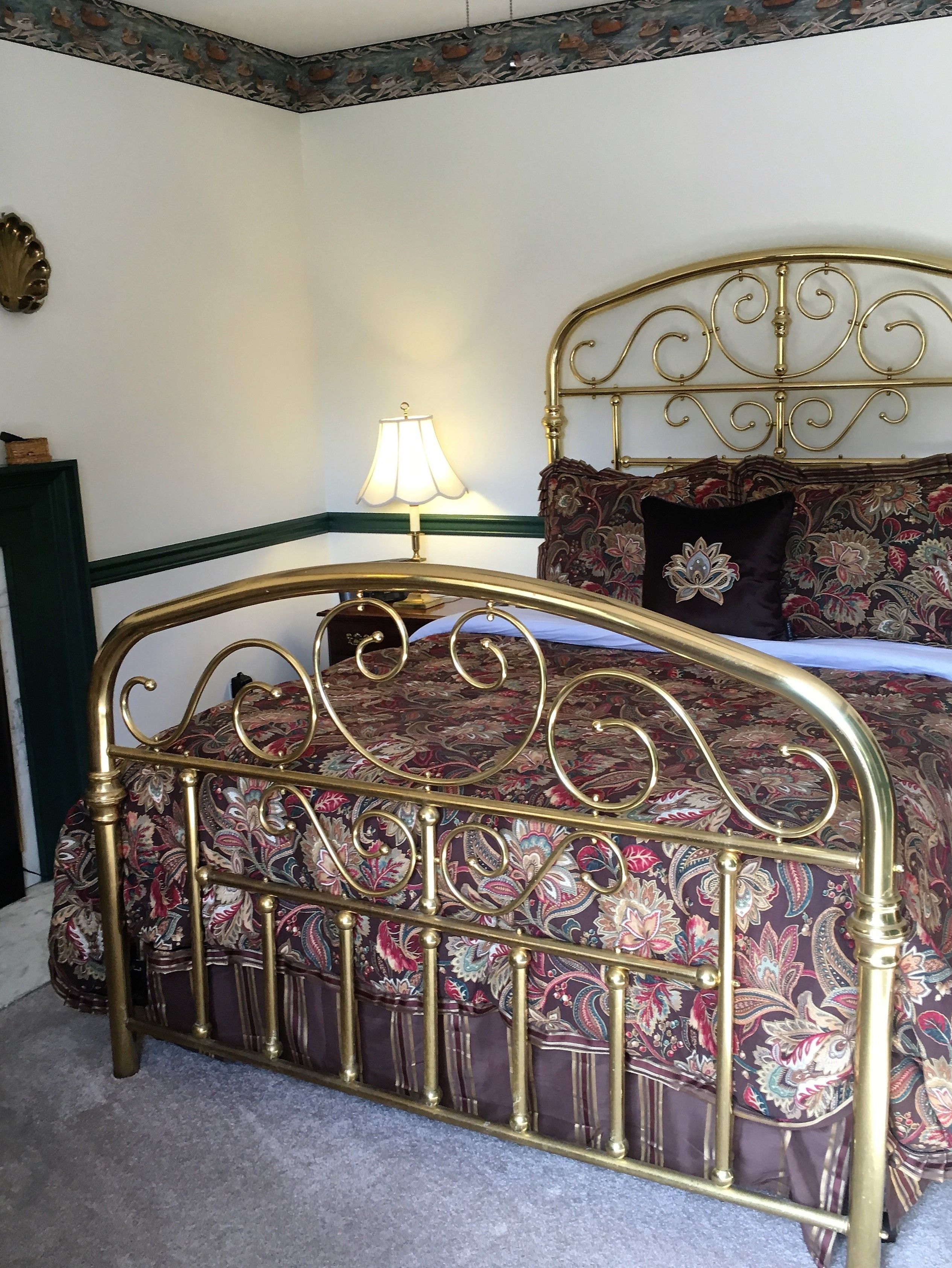 A bed with an ornate metal headboard and a floral-patterned bedspread. A lamp is on the nightstand beside the bed.
