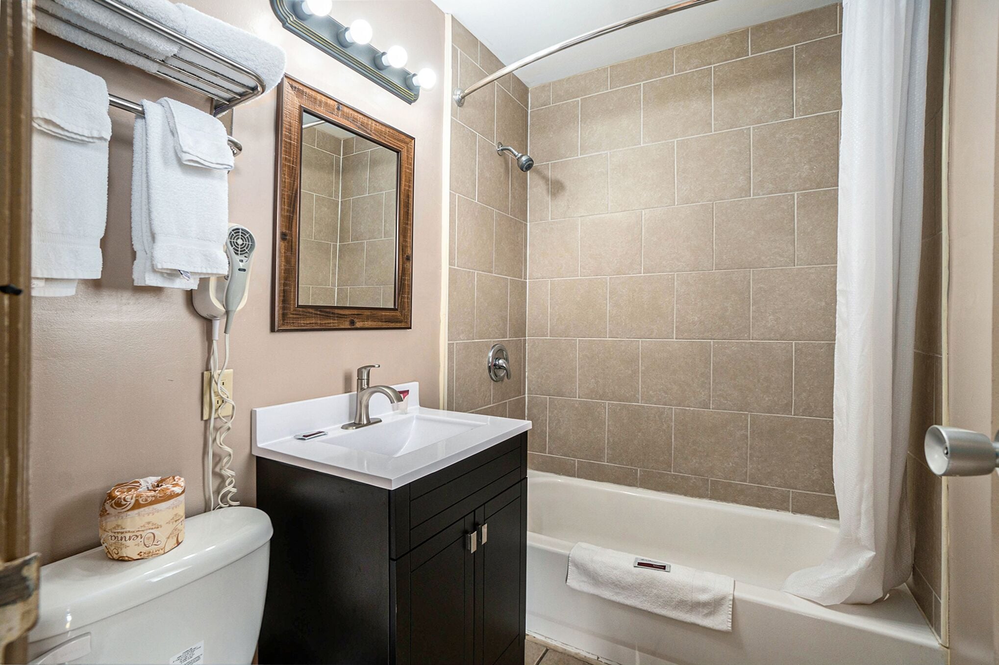 A bathroom with a white sink, dark cabinets, a mirror, a towel rack, and a shower with beige tiles.