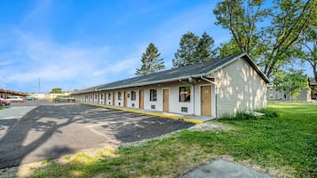 Un motel con una hilera de habitaciones de una sola planta, un estacionamiento y un área verde con árboles.