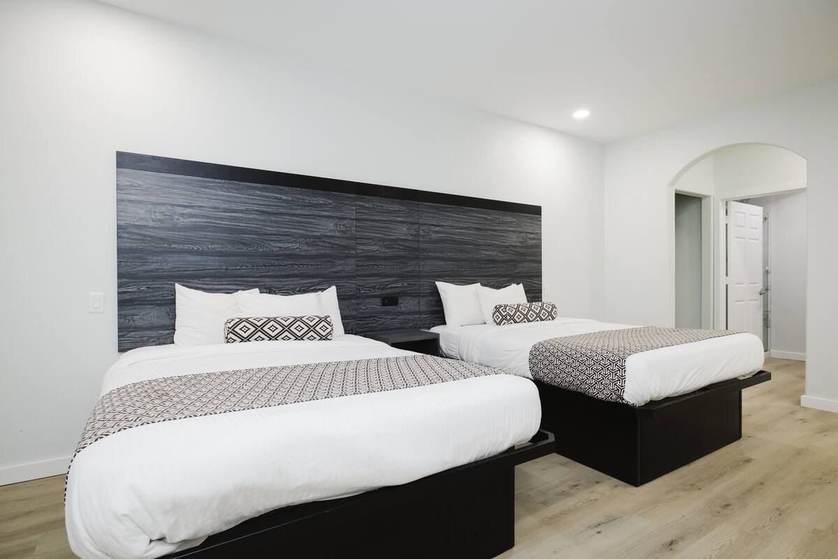A modern hotel room with two beds, a dark wood headboard, white bedding, and a wooden floor.