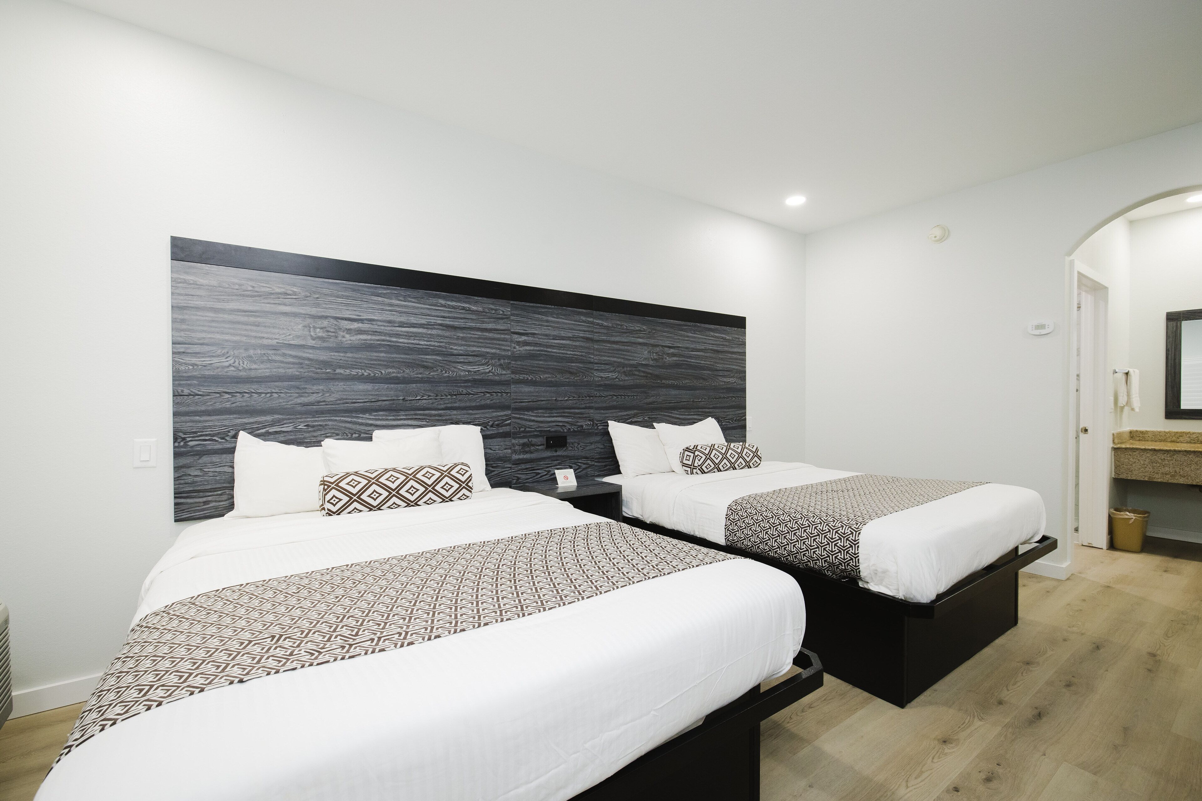 A hotel room with two beds, a dark wood headboard, white bedding, and a small bedside table with a lamp.