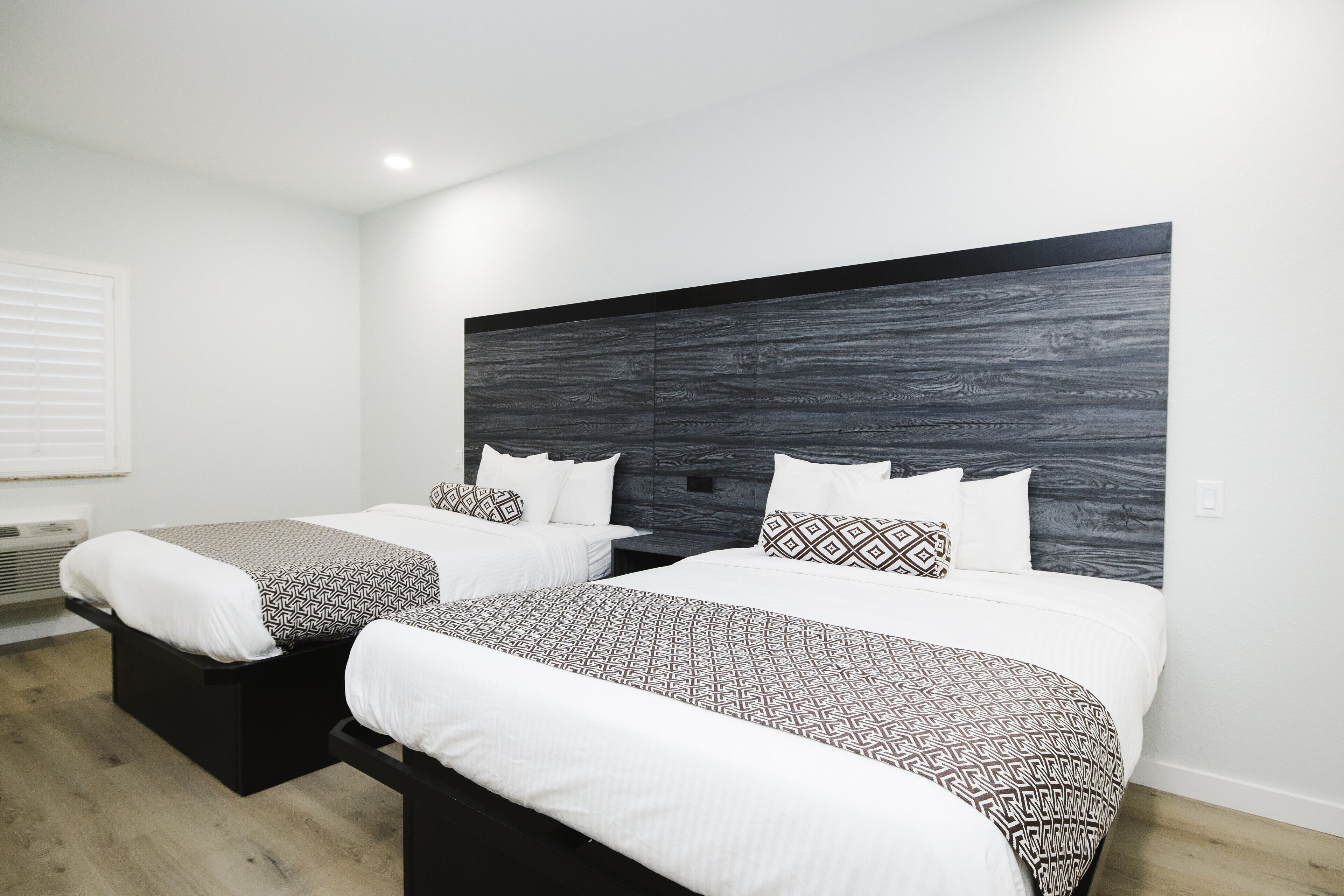 A modern hotel room with two beds, a dark wood headboard, and a white wall.