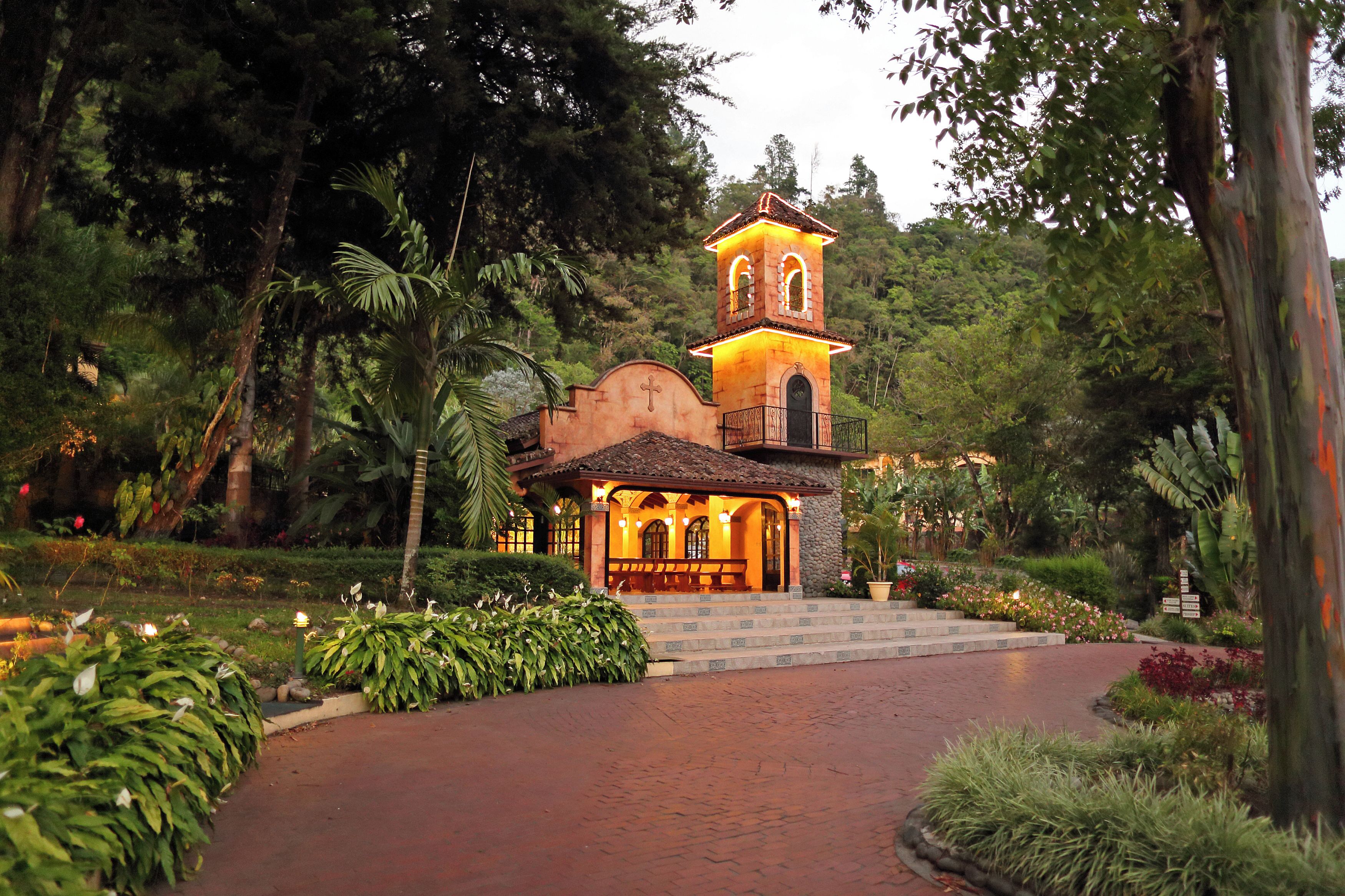 A small, illuminated church with a bell tower, surrounded by lush greenery and a paved pathway leading to it.