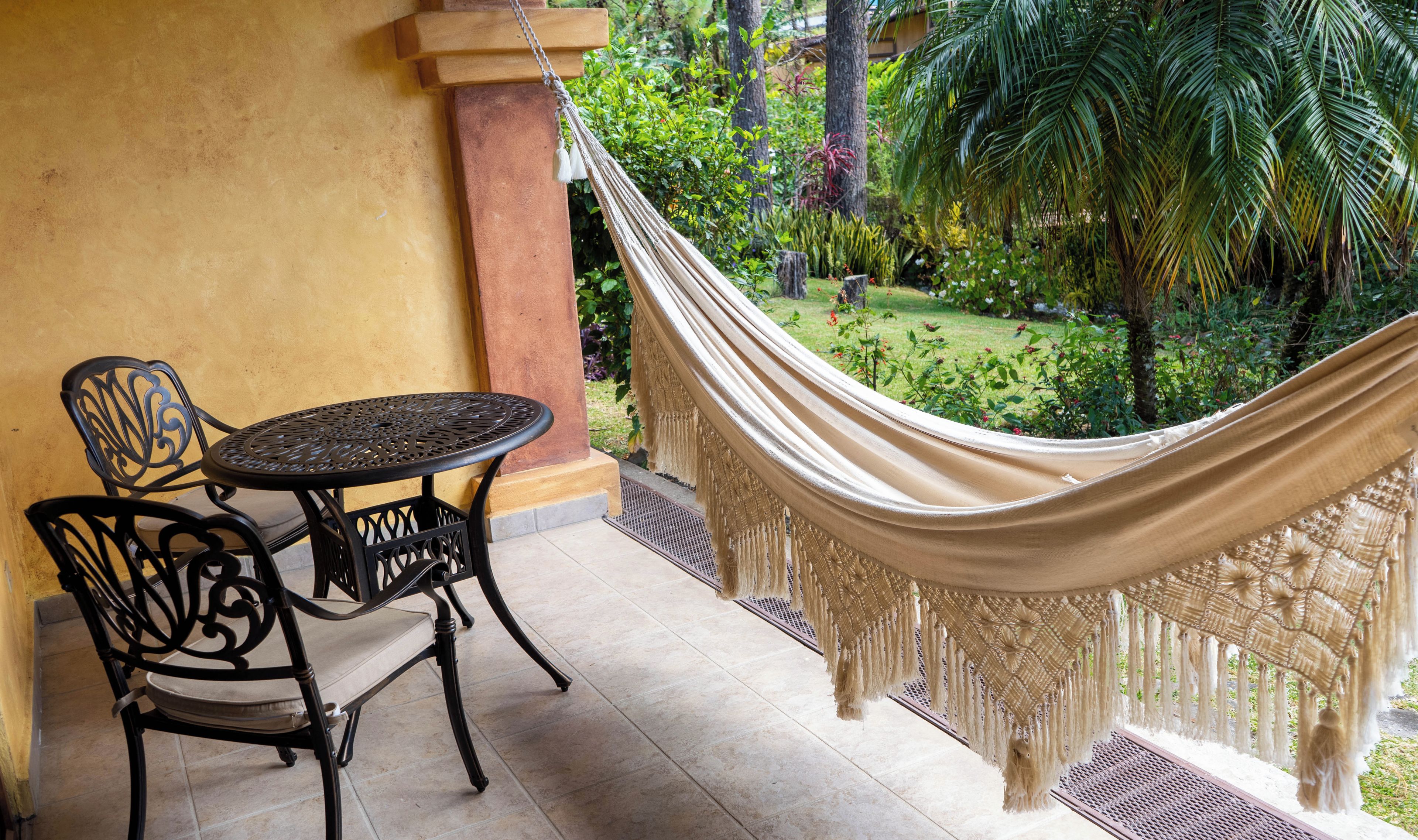 A hammock tied to a column, a round metal table with a chair, and a view of a garden with trees and plants.