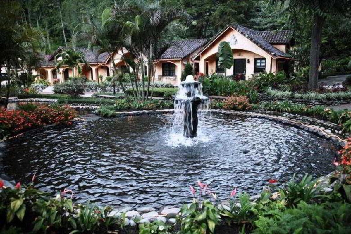 A garden with a central water feature, surrounded by tropical plants and a building with a tiled roof.