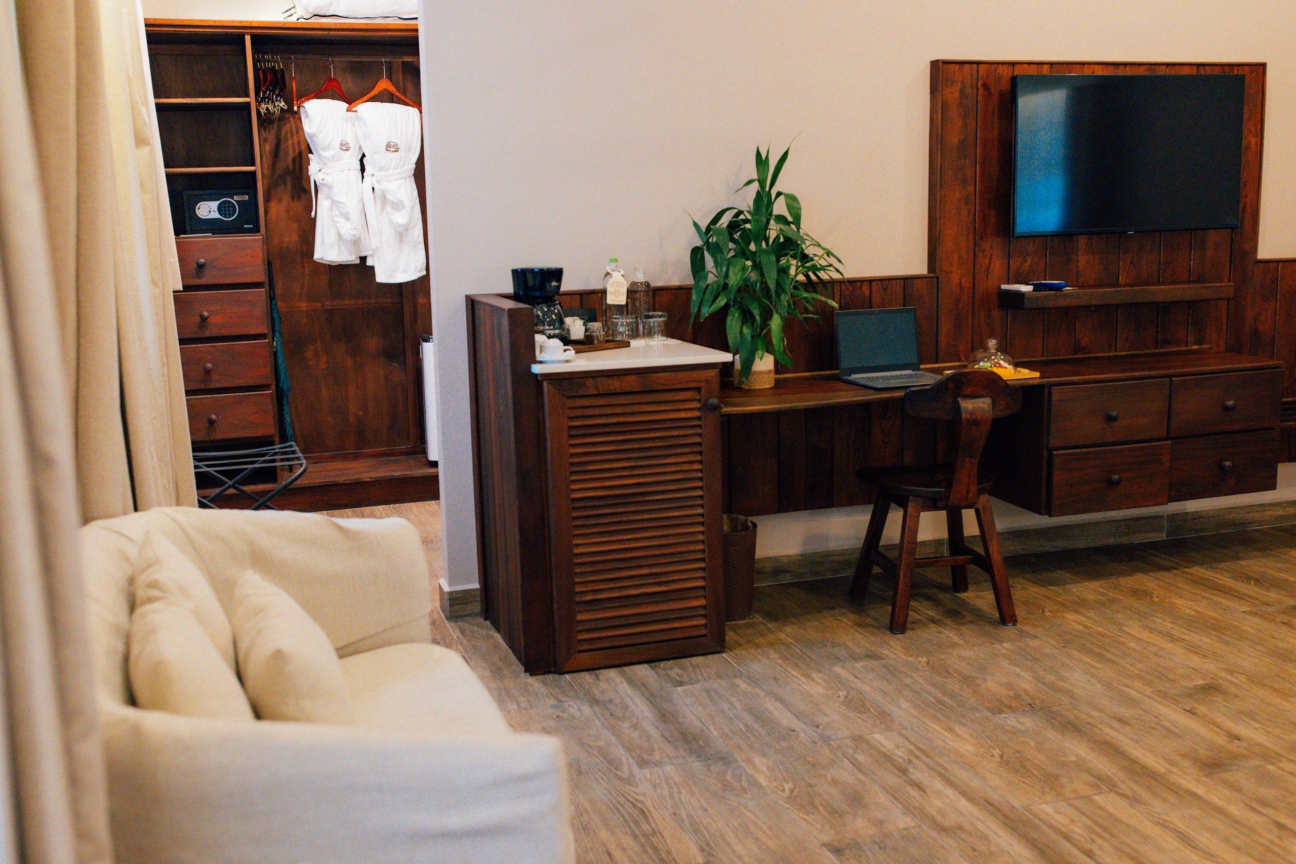 A hotel room with a wooden desk, a chair, a television mounted on the wall, a plant on the desk, and a wardrobe with hanging robes.