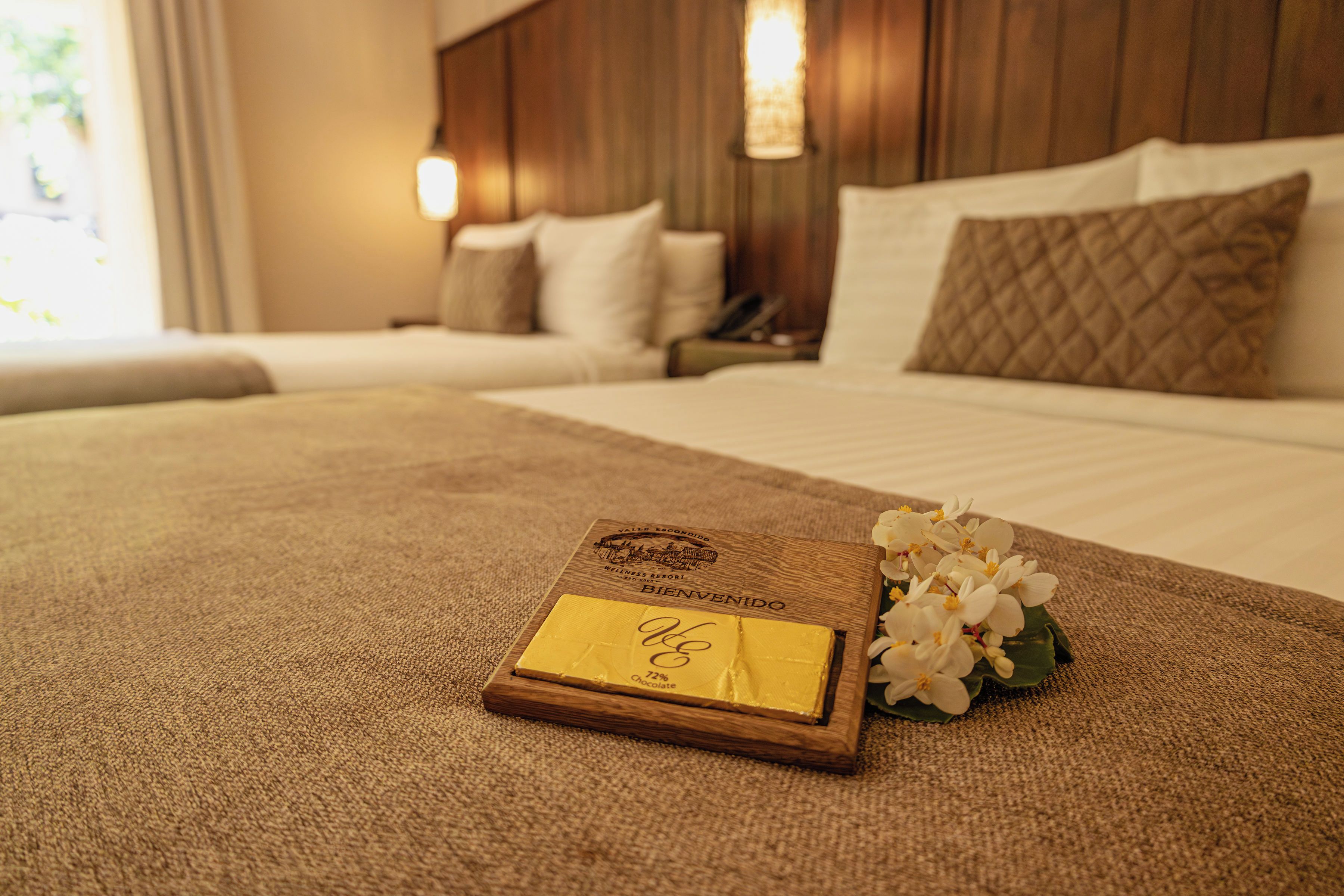 A neatly made bed with a wooden headboard, a bedside table with a lamp, and a welcome card on the bed.