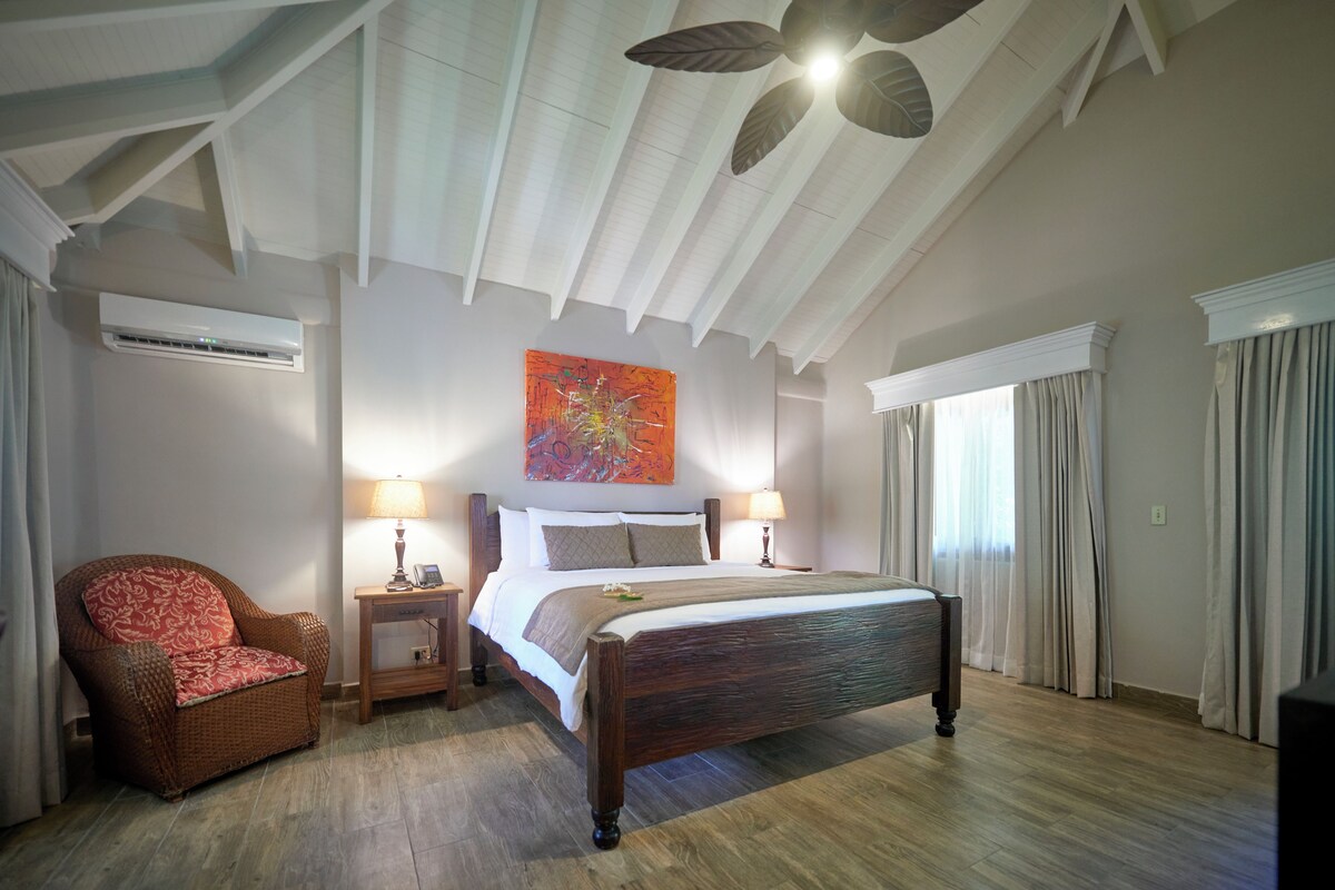 A bedroom with a wooden bed, a wicker chair, a nightstand with a lamp, and a ceiling fan.