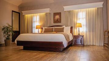 A large bed with a wooden frame, two bedside tables with lamps, a potted plant, and a painting on the wall.