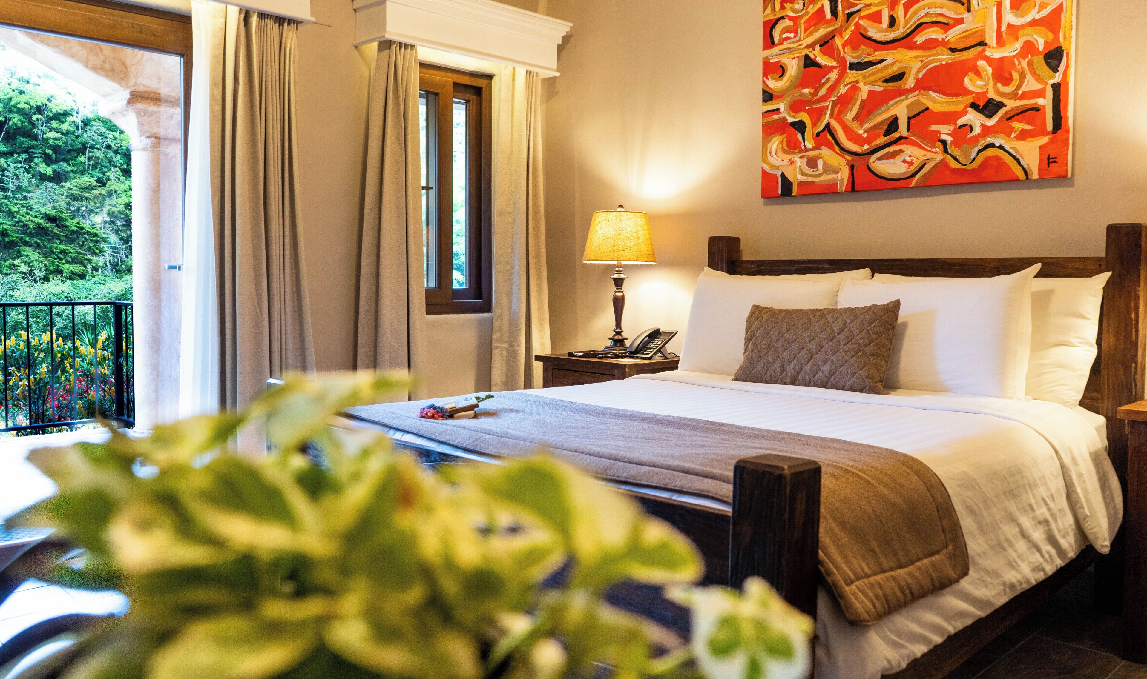 Een hotelkamer met een bed, nachtkastje met lamp, telefoon, raam met gordijnen en een schilderij aan de muur.