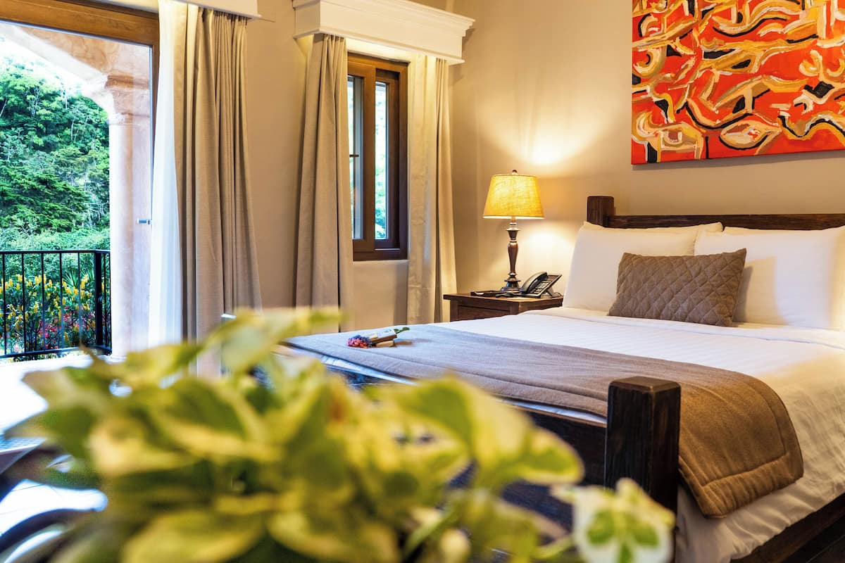 Une chambre d’hôtel avec un lit, une table de chevet avec une lampe, un téléphone, une fenêtre avec des rideaux et un tableau au mur.