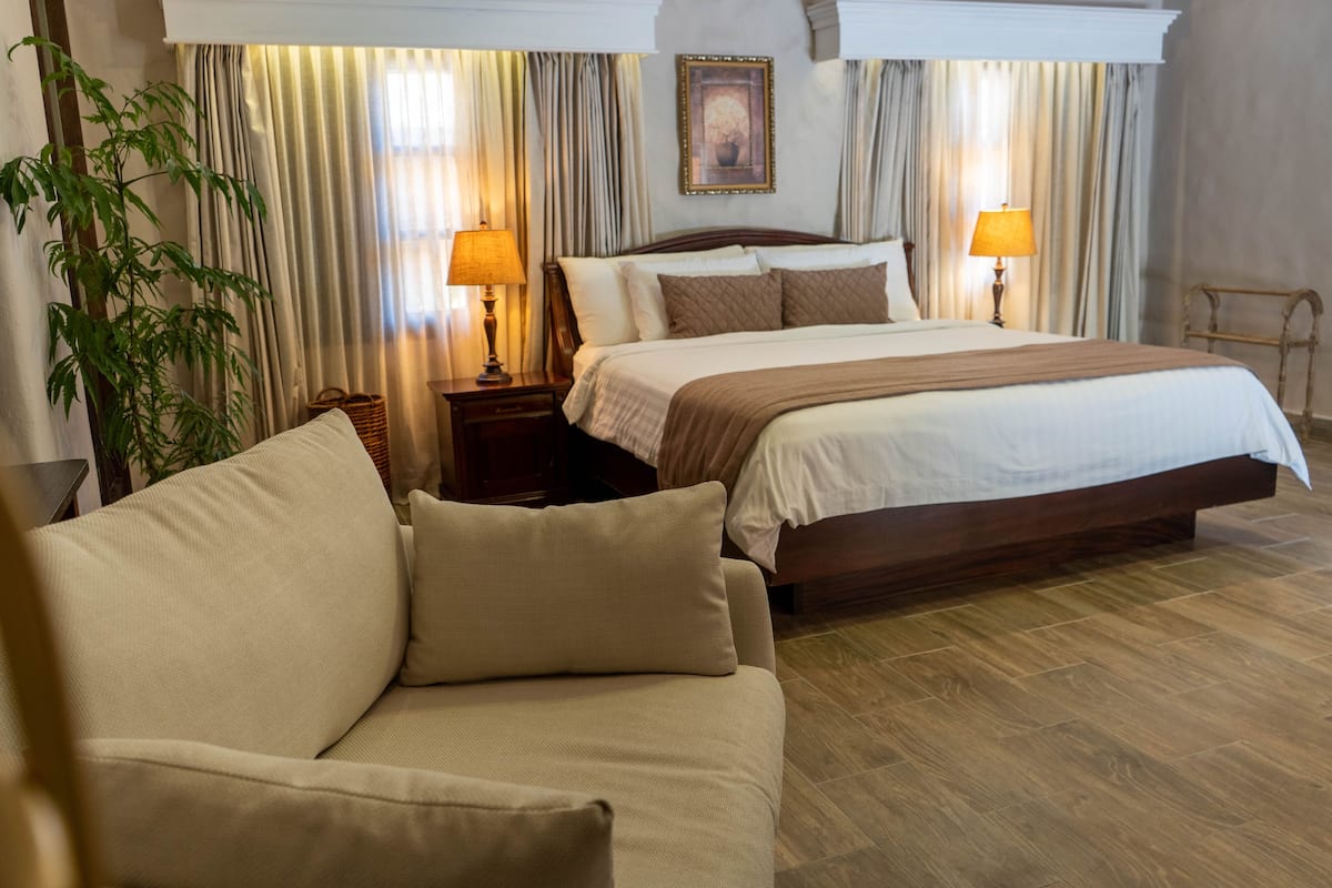 A hotel room with a large bed, two bedside lamps, a wooden headboard, a beige sofa, and a large potted plant.