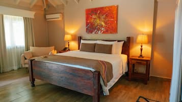 A hotel room with a large bed, two bedside tables with lamps, a chair, a painting on the wall, and an air conditioning unit.