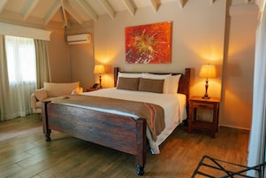 Habitación de hotel con una cama grande, dos mesitas de noche con lámparas, una silla, un cuadro en la pared y un equipo de aire acondicionado.