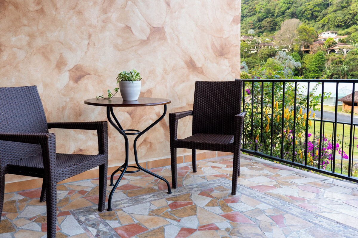 A balcony with two wicker chairs and a small table, a potted plant, and a view of a lush garden and houses in the background.