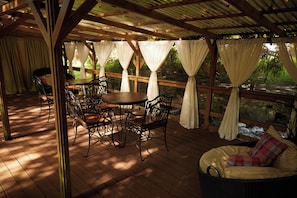 Une terrasse extérieure couverte, dotée d’un plancher en bois, de chaises en métal, d’une table ronde et de rideaux blancs.