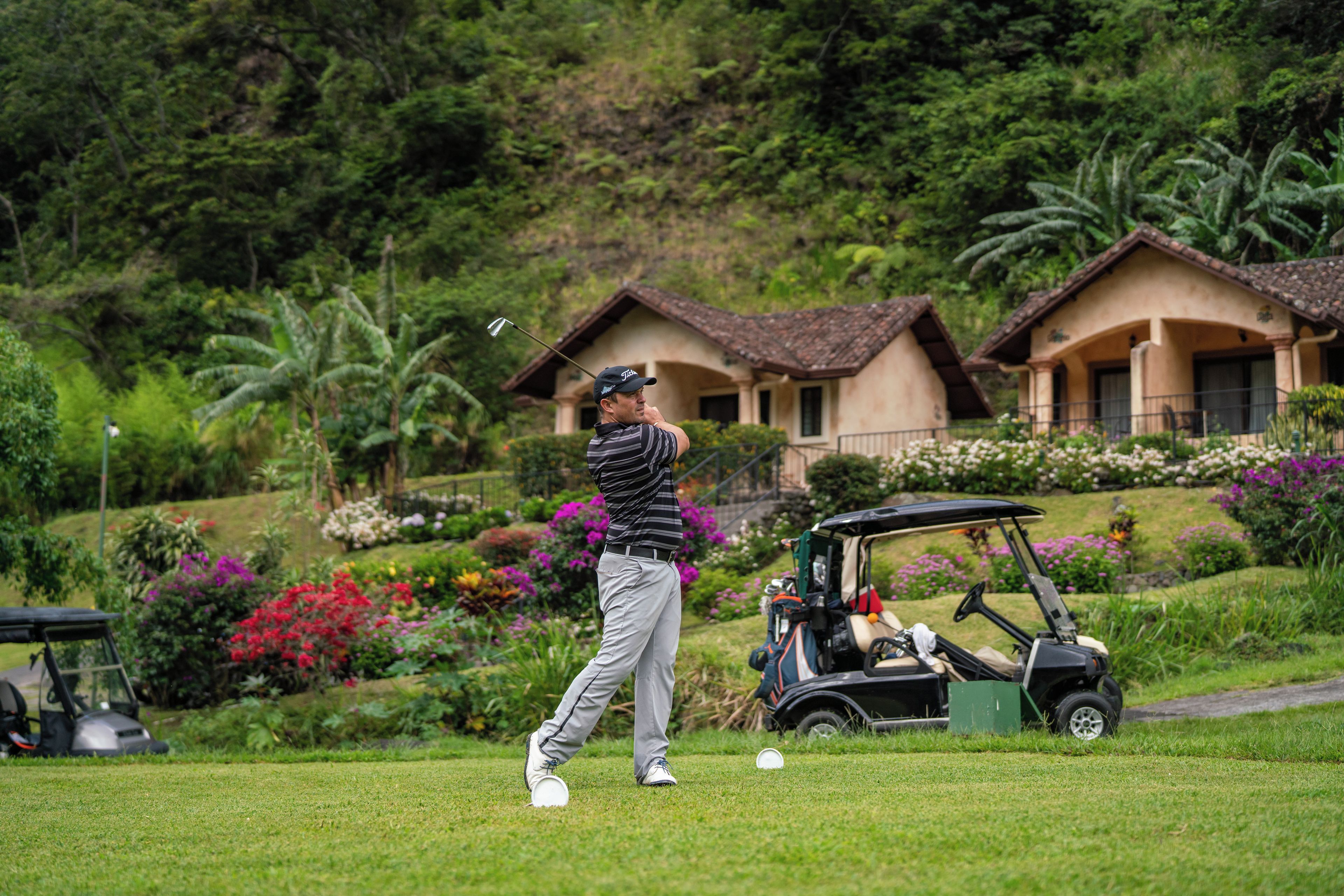 A golf course with a player in mid-swing, a golf cart, and a building with a tiled roof in the background.