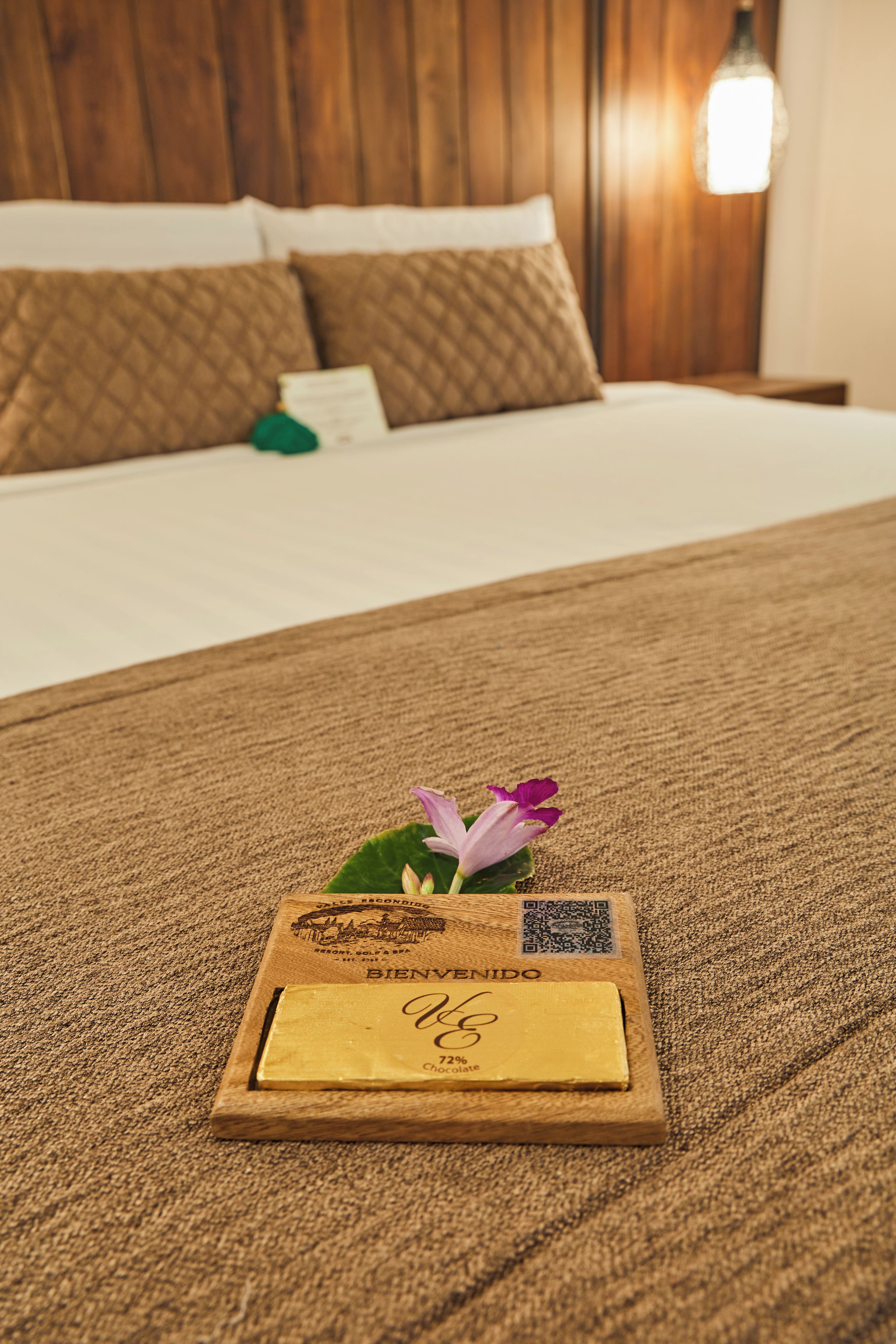 Una habitación de hotel con una cama bien hecha, una mesita de noche de madera con una barra de chocolate y una tarjeta de bienvenida, y un cojín con un detalle en verde.