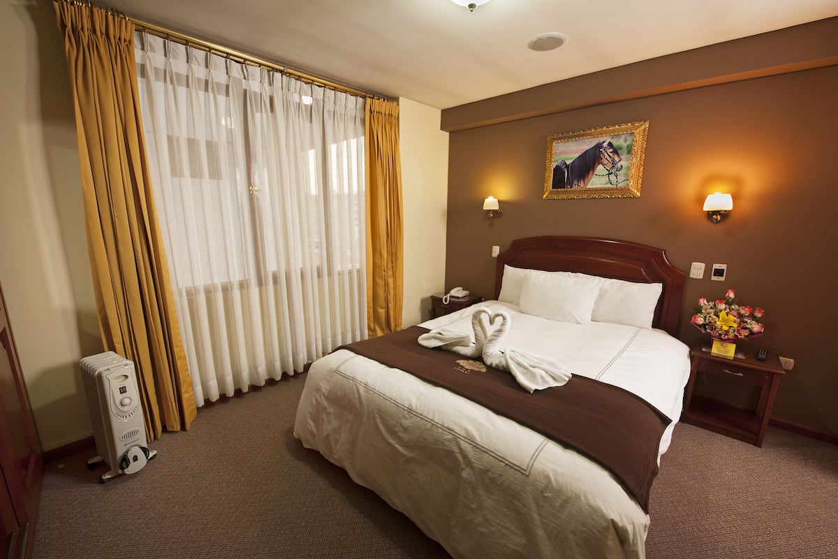 A hotel room with a large bed, a nightstand with a lamp, a painting on the wall, and a window with curtains.