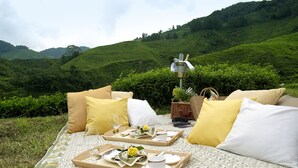 A picnic setup with a blanket, cushions, and trays of tea and snacks on a grassy area with a tea plantation in the background. - Cameron Highlands Resort (Tanah Rata)