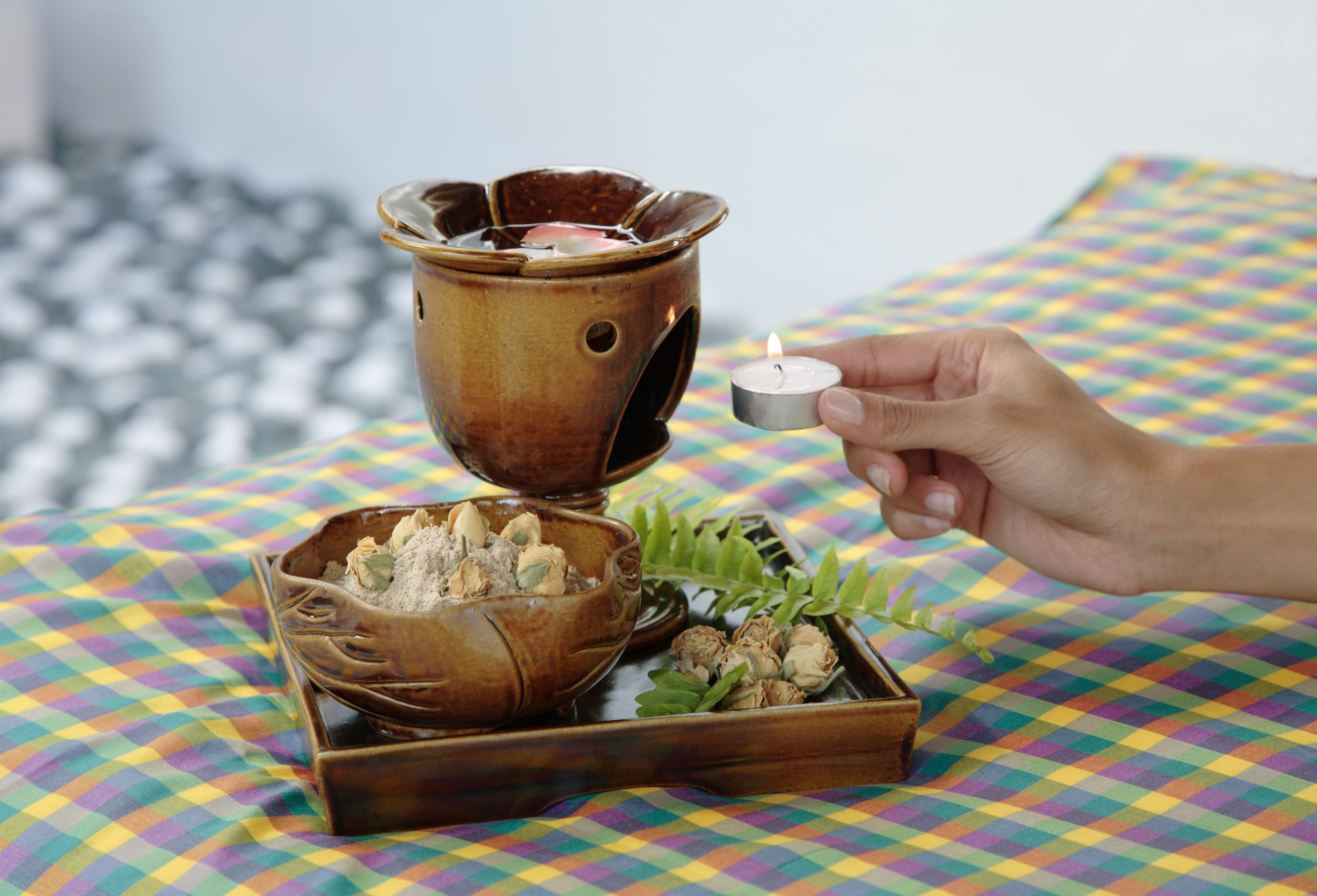 A hand holding a small candle next to a decorative ceramic bowl and a tray with dried flowers on a colorful checkered fabric.