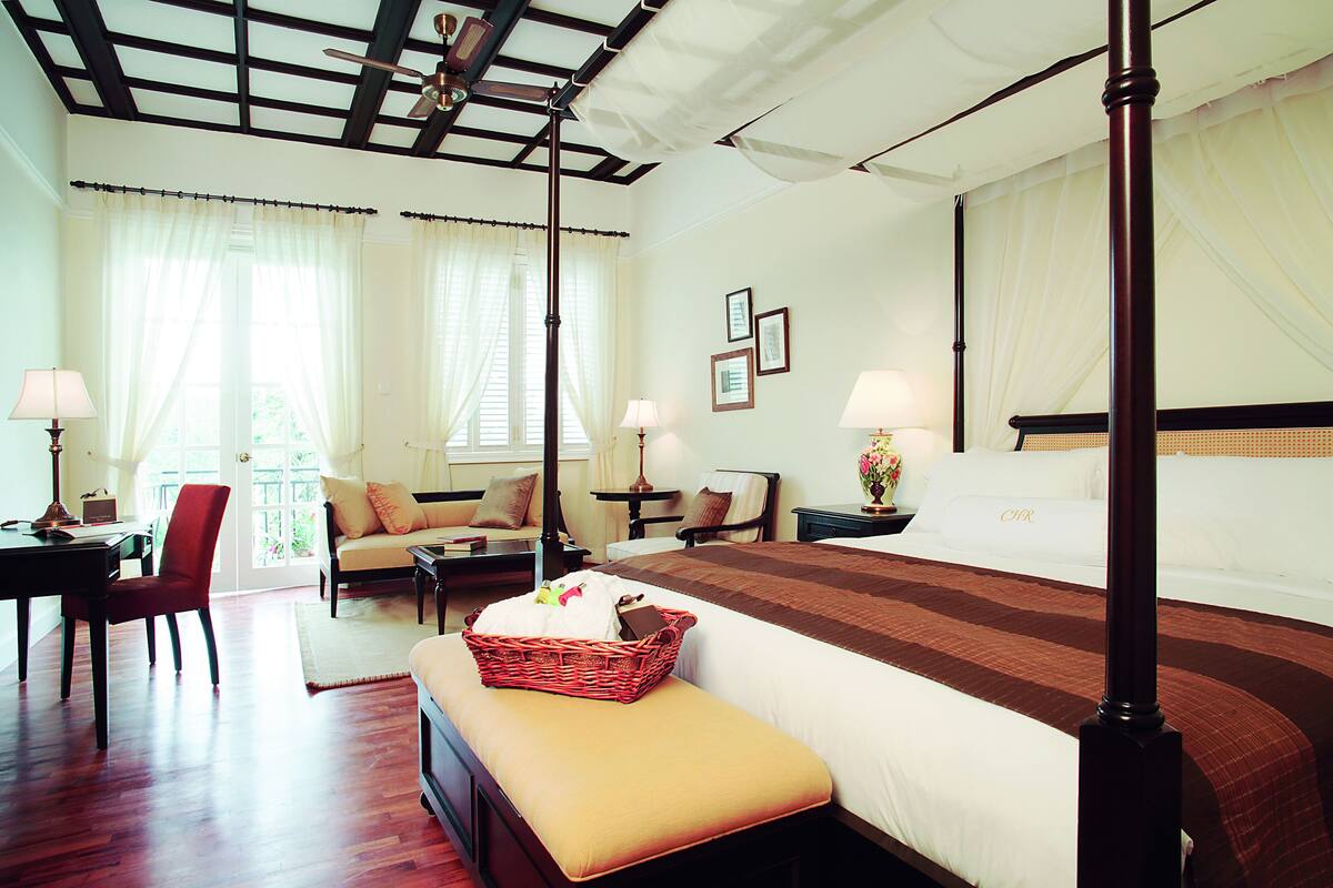 A hotel room with a large bed, a sitting area with a sofa and armchairs, a dining table, and a red wicker basket on the bed.