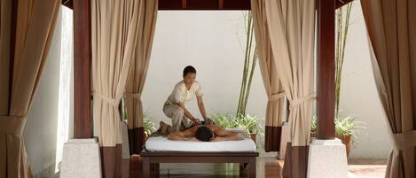 A massage therapy session in a room with a wooden bench, beige curtains, and potted plants.