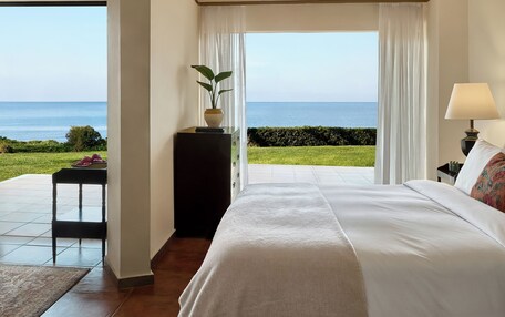 A bedroom with a large bed, a bedside table with a lamp, a potted plant, and a view of the sea.