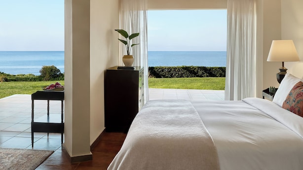 Une chambre avec un grand lit, une table de chevet avec une lampe, une plante en pot et une vue sur la mer.