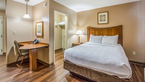 A hotel room with a wooden desk, a bed with white bedding, a wooden headboard, a bedside lamp, and a framed picture on the wall. - MainStay Suites Port Saint Joe South (Port St. Joe)