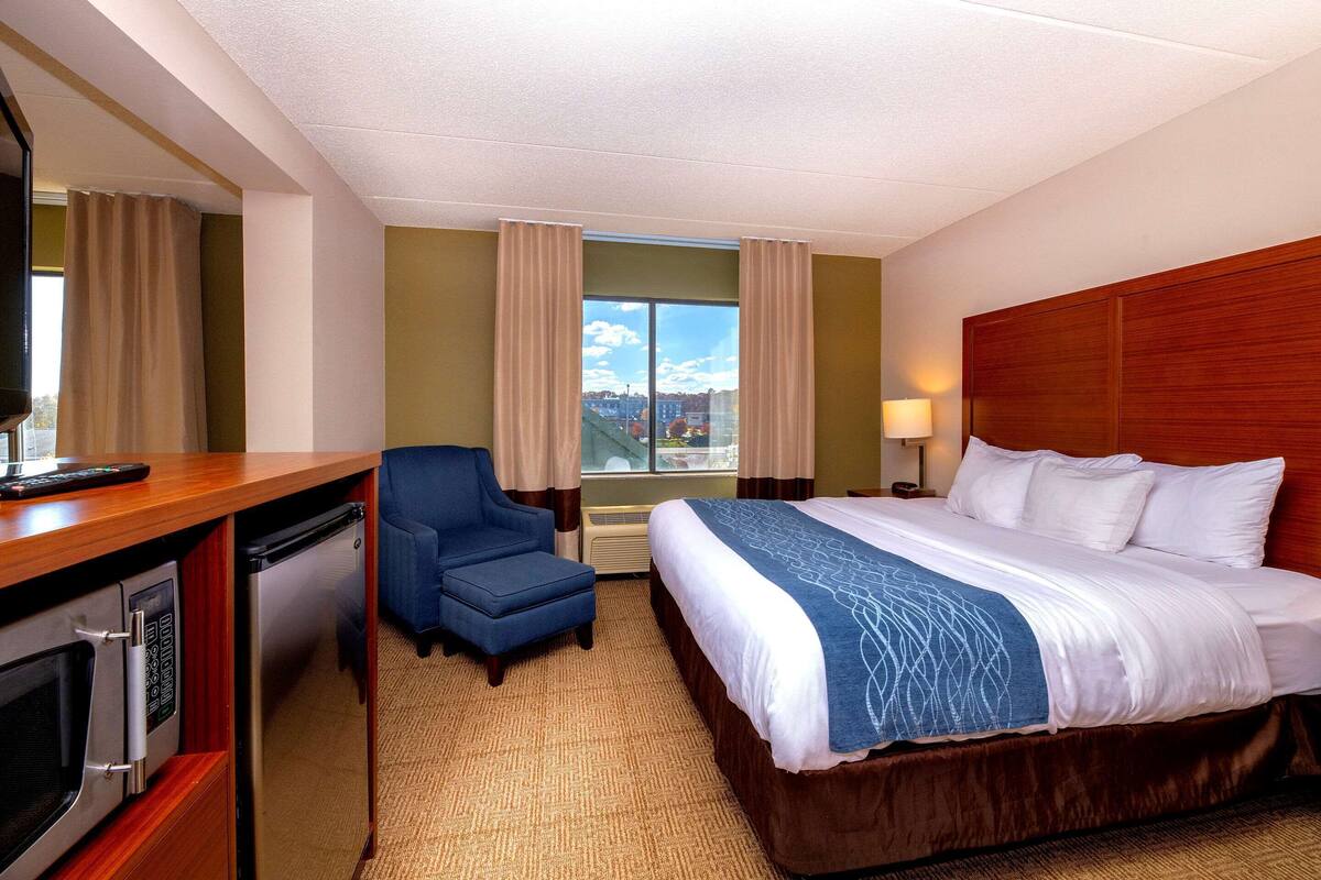 A hotel room with a bed, a desk with a microwave, a chair, and a window with curtains.