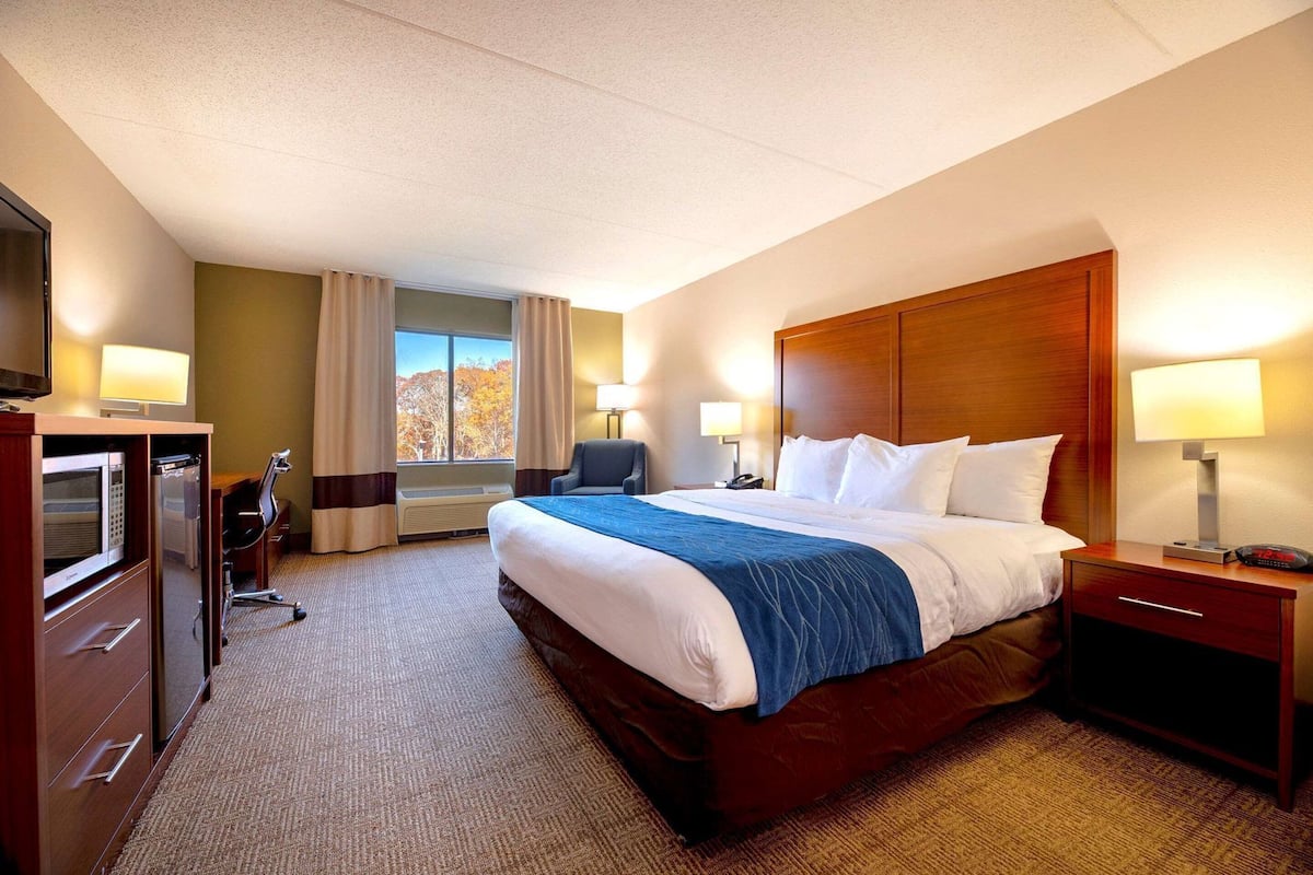 A hotel room with a large bed, a desk with a microwave, a chair, a desk lamp, and a nightstand with a lamp.