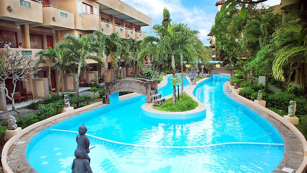 A swimming pool with a statue in the center, surrounded by palm trees and a multi-story building with balconies. - Melasti Beach Resort & Spa (Legian)