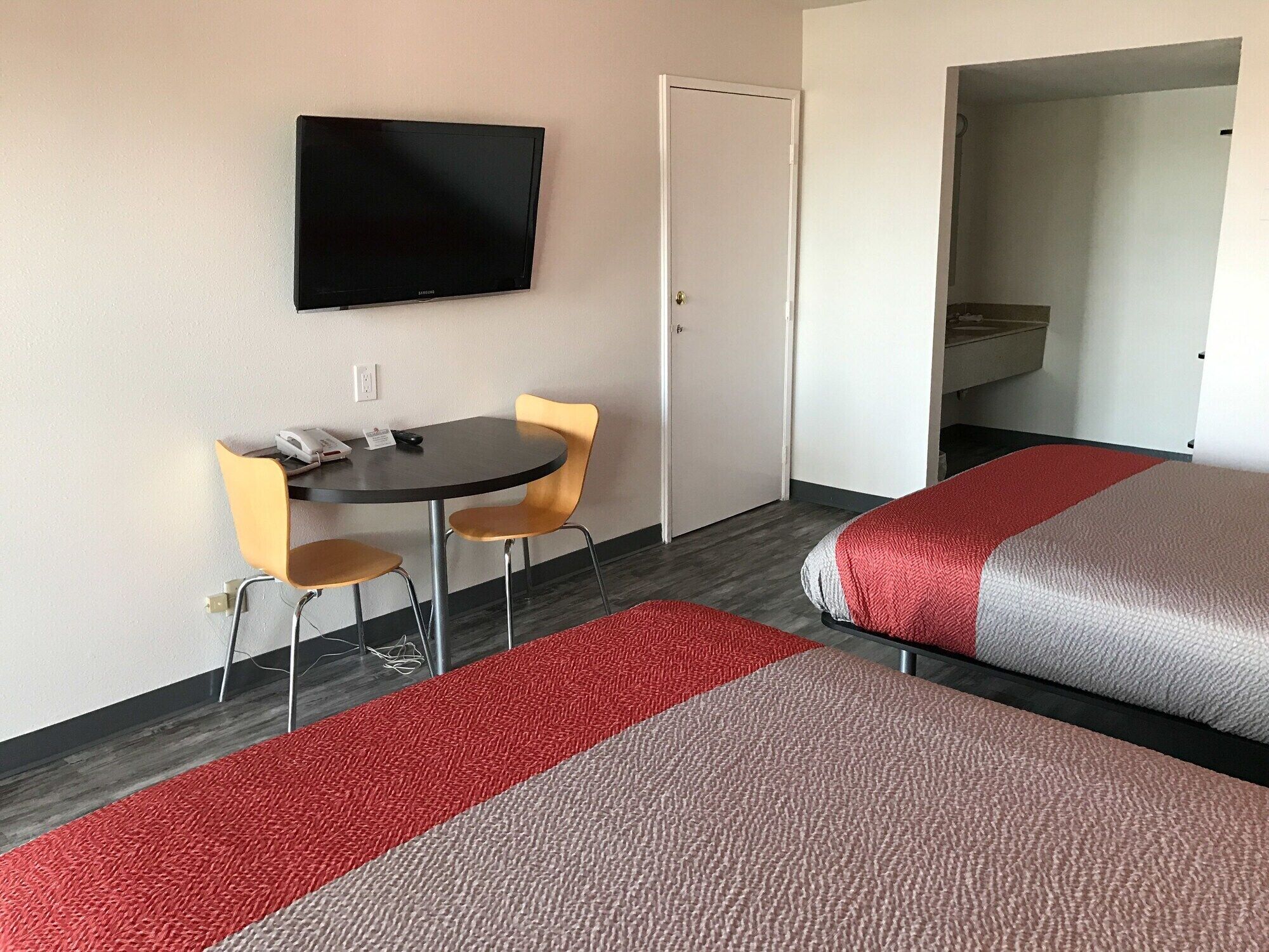 A hotel room with a bed, a TV mounted on the wall, a small table with two chairs, and a door.