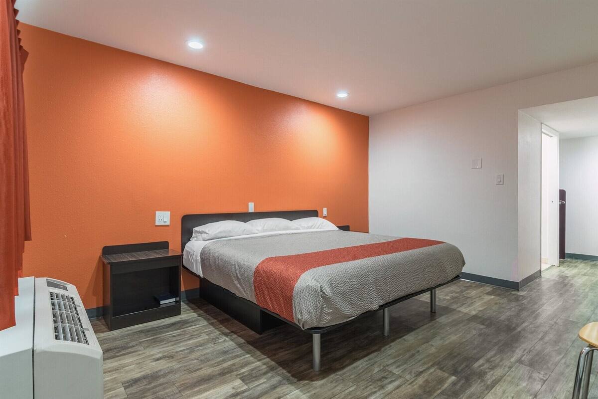 A hotel room with an orange accent wall, a bed with a grey and red blanket, a nightstand with a phone, and a wooden floor.